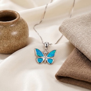 May include: A silver butterfly pendant necklace with a blue and green opal-like design. The butterfly has a small diamond accent. The necklace is displayed on a white satin surface with a small brown vase and a folded brown cloth in the background.