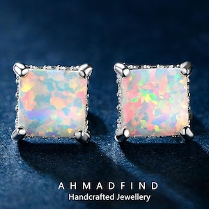 May include: A pair of square stud earrings with iridescent opal centers and silver-colored metal settings. The earrings have a four-prong design and are set against a dark blue background. The text "AHMADFIND Handcrafted Jewellery" is at the bottom.