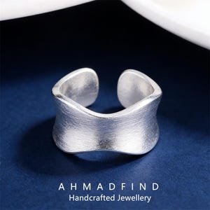 Może przedstawiać: Srebrny pierścionek z wykończeniem ze szczotkowanego metalu. Pierścionek ma unikalny, otwarty design z zakrzywionym, szerokim pasmem. Tekst "AHMADFIND Handcrafted Jewellery" znajduje się na dole obrazu.