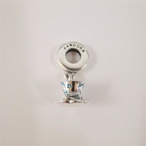 May include: A silver Pandora charm shaped like a teacup and saucer. The teacup features light blue painted floral accents and a small star detail. The charm includes a circular bail at the top with the brand name Pandora engraved in black lettering.