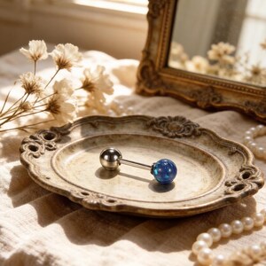 Könnte beinhalten: Ein silberner Piercingschmuck im Hantelstil mit einem blauen Opalball und einem silbernen Ball auf einem dekorativen Teller. Der Schmuck wird auf einem Teller im Vintage-Stil mit einer Perlenkette und einem goldgerahmten Spiegel im Hintergrund präsentiert.