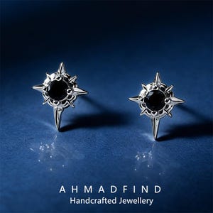 May include: A pair of silver-colored stud earrings. Each earring features a black gemstone center surrounded by a starburst design. The earrings are on a blue background. The text "AHMADFIND Handcrafted Jewellery" is at the bottom.