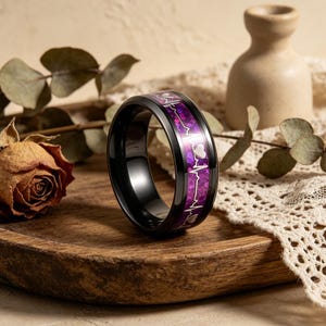Könnte beinhalten: Ein schwarzer Ring mit einem lila Band mit einem Herzschlag-Design und kleinen Herzakzenten. Der Ring liegt auf einem Holztablett mit einer getrockneten Rose und dekorativen Elementen im Hintergrund.