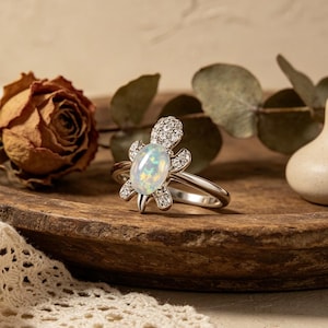 May include: A silver ring featuring a turtle design. The turtle's body is an oval opal, and the head and flippers are adorned with small, clear gemstones. The ring is set on a wooden surface with a dried rose and decorative elements.