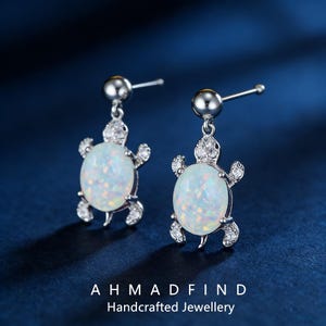 Könnte beinhalten: Silberfarbene Ohrringe in Schildkrötenform mit weißen Opal-Effekt-Steinen und klaren Kristallakzenten. Die Ohrringe hängen an silbernen Stiften. Der Text "AHMADFIND Handcrafted Jewellery" ist unten im Bild zu sehen.