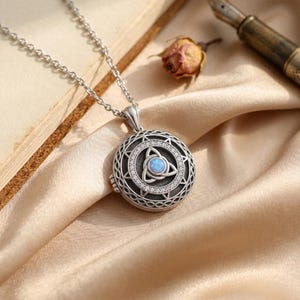 May include: Silver-toned pendant necklace with a round locket design. The locket features a Celtic knot design with a central blue opal stone and is adorned with small clear crystals. The necklace rests on a satin fabric.