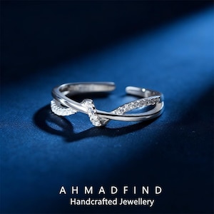 May include: A silver-colored ring with a knot design and small, clear gemstones. The ring has a double-banded design with a crossover knot detail. The ring is displayed on a dark blue surface. The text "AHMADFIND Handcrafted Jewellery" is at the bottom.