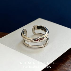 May include: A silver-colored, open-ended ring with a two-tiered design. The top tier has an organic, flowing shape with cut-out details. The bottom tier is a smooth, curved band. The ring is displayed on a white surface with the text "AHMADFIND Handcrafted Jewellery".
