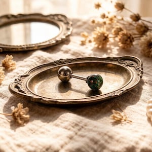 May include: A silver barbell-style body piercing jewelry with a spherical end and a green and blue marbled end, resting on a vintage-style metal tray. The tray has an ornate, scalloped edge. A small, round mirror is in the background.