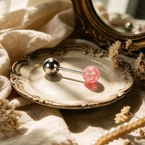 Könnte beinhalten: Ein Piercing im Barbell-Stil mit einer silbernen Metallkugel und einer rosa, glitzernden Kugel. Der Schmuck liegt auf einem dekorativen, cremefarbenen Teller mit goldenem Rand. Natürliches Licht beleuchtet das Bild.