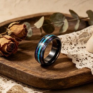 May include: A black ring with a colorful inlay of blue, purple, and white bands. The ring is set on a wooden tray with dried roses and greenery. The ring's design is eye-catching and modern.