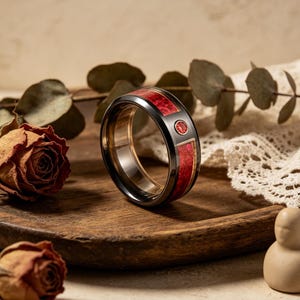 Könnte beinhalten: Ein schwarzer Ring mit roten und goldenen Einlagen. Der Ring hat einen kreisförmigen roten Stein, der in einen rechteckigen goldenen Rahmen gefasst ist. Der Ring wird auf einer Holzoberfläche mit getrockneten Rosen und Grünzeug präsentiert.