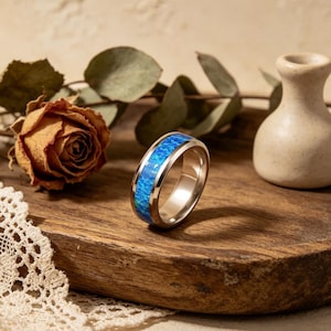 Könnte beinhalten: Ein silberner Ring mit einem lebendigen blauen Opal-Inlay. Der Ring liegt auf einer Holzoberfläche, mit einer getrockneten Rose und einer kleinen Vase im Hintergrund. Der Opal hat ein helles, gesprenkeltes Aussehen.