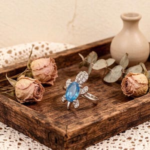 May include: A silver ring with a turtle design, featuring a large oval, light blue gemstone as the shell. The turtle's head and flippers are decorated with small, clear gemstones. The ring is displayed on a wooden tray with dried roses and a small vase.