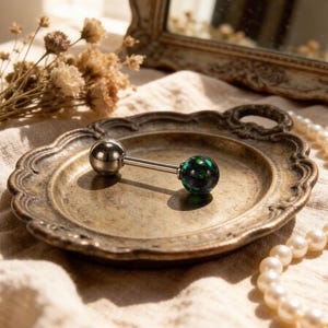 Könnte beinhalten: Ein silberner Zungenpiercing-Barbell mit einer metallischen Kugel und einer dunkelgrünen, irisierenden Kugel. Der Schmuck wird auf einem antiken, verzierten, bronzefarbenen Teller präsentiert.