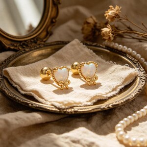 May include: Gold-coloured heart-shaped earrings with a white opal centre and a bow detail. The earrings are displayed on a gold-rimmed plate with a beige cloth. A gold-framed mirror and pearls are in the background.