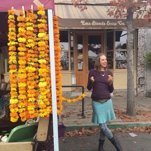May include: A display of long, vertical garlands made of bright orange and yellow marigold flowers hanging from a frame. Beside the garlands, a person wears a long-sleeved dark purple shirt, a light blue skirt, and patterned grey leggings.