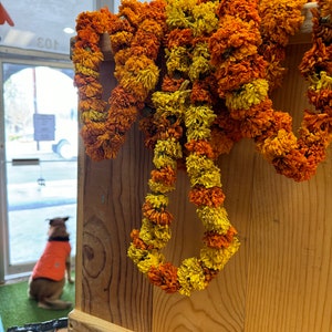 May include: A decorative garland made of dried marigold flowers in vibrant shades of orange and yellow, draped over a light-colored wooden crate. The flowers are densely packed, creating a textured, layered appearance across the wooden surface.