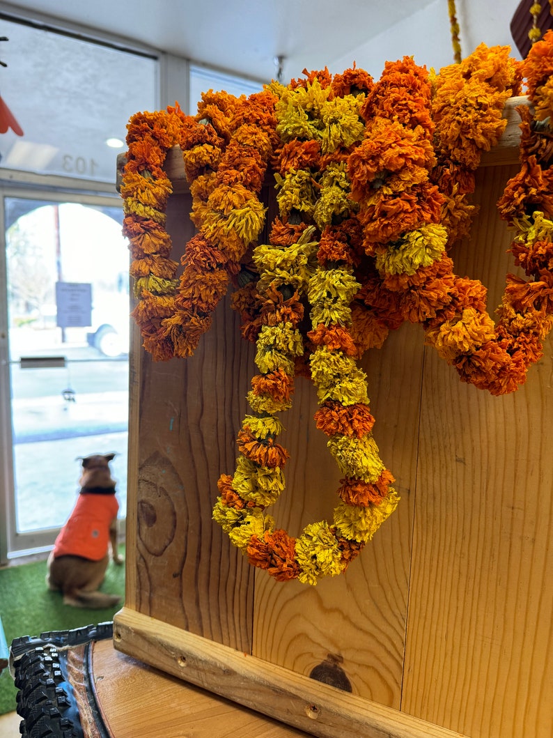 May include: A decorative garland made of dried marigold flowers in vibrant shades of orange and yellow, draped over a light-colored wooden crate. The flowers are densely packed, creating a textured, layered appearance across the wooden surface.