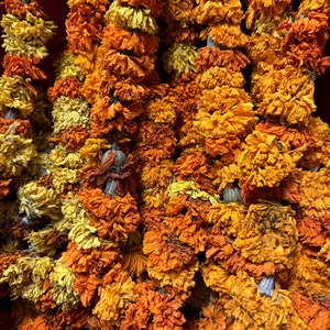 May include: Multiple vertical strands of dried marigold flowers in shades of deep orange and bright yellow. The individual blossoms are densely packed along each string, showing a textured, crinkled appearance characteristic of dehydrated petals.
