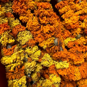 May include: A dense arrangement of dried marigold flowers strung together in long garlands. The petals display a vibrant spectrum of deep orange, burnt orange, and bright yellow hues, creating a textured, layered appearance throughout the composition.