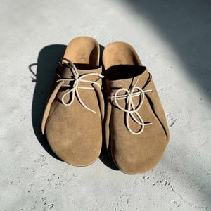 Può includere: Un paio di zoccoli in pelle scamosciata marrone chiaro con lacci bianchi annodati a fiocco. Gli zoccoli hanno una punta arrotondata e un tacco basso. Le scarpe sono su una superficie grigio chiaro.
