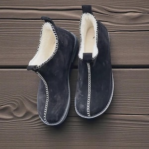 May include: A pair of black suede slippers with white stitching and a soft, white interior lining. The slippers have a pull tab at the back and are designed for indoor wear. The soles are a dark gray color.