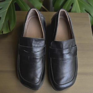 May include: A pair of black leather loafers with a classic penny strap design. The shoes have a rounded toe and a light brown interior. The loafers are displayed on a wooden surface, with green foliage in the background.