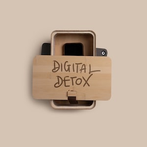 May include: A light brown bamboo box with the words "DIGITAL DETOX" written on the lid. Inside the box is a black smartphone. The box is open, revealing the phone and the interior.