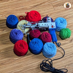 May include: A rug tufting gun with a blue handle is surrounded by colorful yarn balls in shades of red, blue, green, and purple. The tool is silver and black, with a power cord extending from the handle. The logo "Tankard's Tapestries" is in the upper right corner.