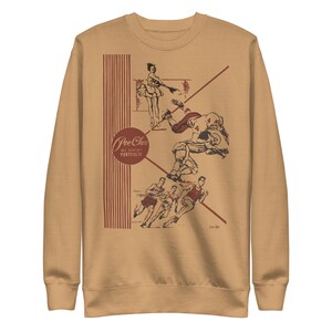 May include: Tan sweatshirt with a vintage-style graphic. The graphic features illustrations of a football player, runners, and a dancer, with the text "Pee Chee All Season Portfolio" in a circular design. The sweatshirt has a crew neck and long sleeves.