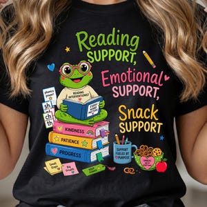 May include: A black t-shirt featuring a green frog wearing pink glasses and reading a book. The text reads Reading SUPPORT, Emotional SUPPORT, Snack SUPPORT. Below are stacked books labeled Kindness, Patience, Progress, and a mug with snacks.