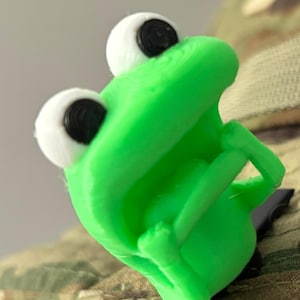 May include: A bright green frog-shaped clip with white and black eyes. The clip is attached to a camouflage fabric. The frog's mouth is open, and it appears to be made of plastic.