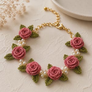 Könnte beinhalten: Ein Armband mit fünf gehäkelten rosa Rosen, kleinen grünen gehäkelten Blättern und Clustern aus weißen Kunstperlen. Die floralen Elemente sind durch eine goldfarbene Metallkette mit Karabinerverschluss und Verlängerungskettchen verbunden.