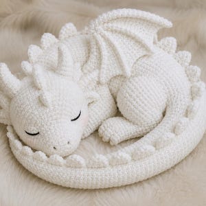 May include: A white crocheted dragon plush toy in a curled sleeping position. The creature features detailed stitched eyes, small horns on its head, a row of spikes along its back, and a single visible wing. The texture is uniform and soft.