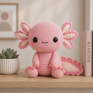 May include: A handmade pink crochet axolotl plush toy with white and pink gills, black safety eyes, and a smiling face. The plush is sitting on a wooden surface. The axolotl is a cute and cuddly handmade item.