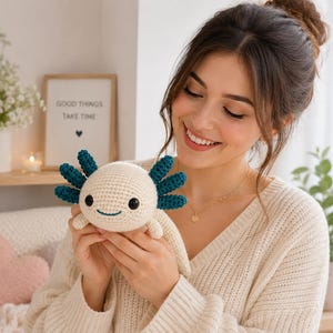 May include: A cream-colored crocheted axolotl plush toy featuring dark teal external gills, black plastic safety eyes, and a stitched teal mouth. The stuffed animal has a rounded head and small limbs, crafted with a visible yarn texture.