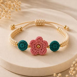 Peut inclure: Un bracelet en macramé avec une fleur crochetée rose au centre, deux accents circulaires crochetés bleu sarcelle et de petites perles dorées. Le bracelet est en cordon crème avec un nœud coulissant réglable et des perles dorées aux extrémités.