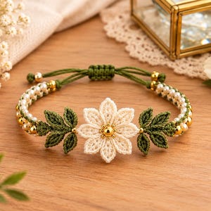 May include: A macrame bracelet featuring a central cream-colored crocheted flower with a gold bead center, flanked by two green crocheted leaves. The band consists of green cord, small white pearls, and gold beads, finished with an adjustable knot.