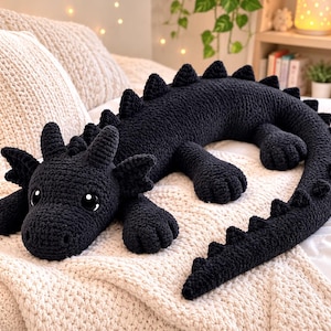 May include: A long, black crochet dragon plush toy featuring a textured yarn surface, large dark eyes with white highlights, small pointed ears, and a series of triangular spikes running along its back and tail, positioned in a lying posture.