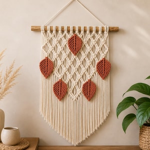 Pode incluir: Uma tapeçaria de macramé feita com corda atada em tom off-white, suspensa por uma haste de madeira. O design inclui cinco detalhes em forma de folha na cor terracota integrados numa rede de losangos, com franjas longas na extremidade inferior.