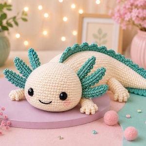 Puede incluir: Un peluche de axolote tejido a ganchillo en color crema con branquias externas y aleta dorsal de color verde azulado. La criatura tiene ojos de plástico negro, una sonrisa bordada y mejillas rosadas, sobre una base redonda de color morado.