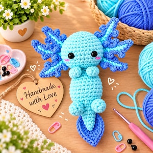Peut inclure: Une peluche axolotl crochetée bleu clair et bleu foncé avec des yeux noirs et des joues roses. À côté se trouve une étiquette en bois en forme de cœur avec le texte "Handmade with Love" imprimé en noir, entouré de petits cœurs roses.