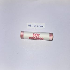 Könnte beinhalten: Eine Rolle mit 50-Cent-Münzen, mit rotem Text "50¢ Pennies" auf einer weißen Papierverpackung. Die Enden der Rolle haben rote Streifen. Eine handschriftliche Notiz über der Rolle lautet "1982 SD Copper".