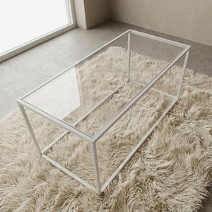 May include: A rectangular coffee table featuring a clear glass top and a white metal frame. The table has a clean, open-base design with four vertical legs connected by horizontal support bars, creating a simple, geometric, and transparent structure.