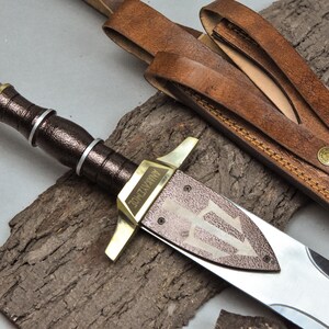 May include: A dagger with a polished brass pommel and crossguard, a dark textured grip with silver rings, and a blade featuring a trident emblem and the text ANAKLYSMOS. It is accompanied by a brown leather sheath with brass rivets and straps.