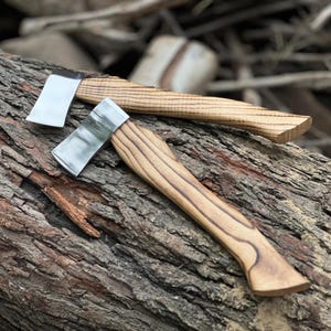 May include: Two small axes with wooden handles and silver blades rest on a weathered log. The handles display a natural wood grain pattern, and the blades have a polished, reflective surface. The axes are designed for outdoor use.