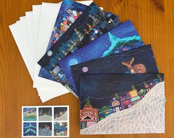 May include: A set of rectangular greeting cards featuring dark blue night sky illustrations with colorful townscapes, pine trees, and a deer, paired with white envelopes and a small sticker sheet displaying six miniature versions of the card designs.