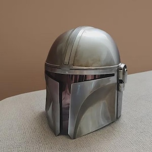 May include: A silver-colored helmet with a rounded top and a visor. The helmet has a metallic finish and is likely a replica or prop. The design includes vertical lines on the top and side details. It is sitting on a light-colored surface.