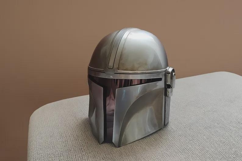 May include: A silver-colored helmet with a rounded top and a visor. The helmet has a metallic finish and is likely a replica or prop. The design includes vertical lines on the top and side details. It is sitting on a light-colored surface.